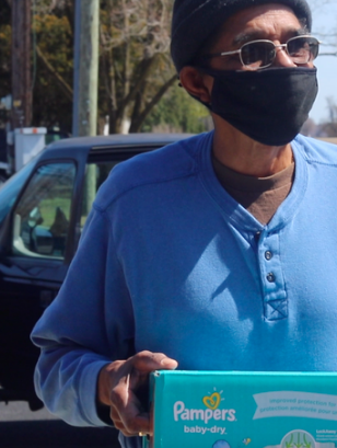 Older man wearing mask, walking, holding box of pampers