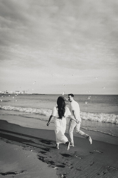 A couple running barefoot on the beach with seagulls flying around them shot in black and white. 