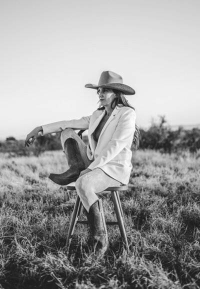 Black and white photo of Juliet Trnka, outdoors sitting on a chair, wearing cowboy boots and hat and a beige blazer. Direct Knowing brand photos