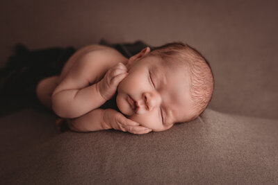 Baby boy at his newborn portrait session at marietta ga newborn photography studio laying on side asleep, wrapped in white white laying on white fabric with hands clasped by his chin and smiling