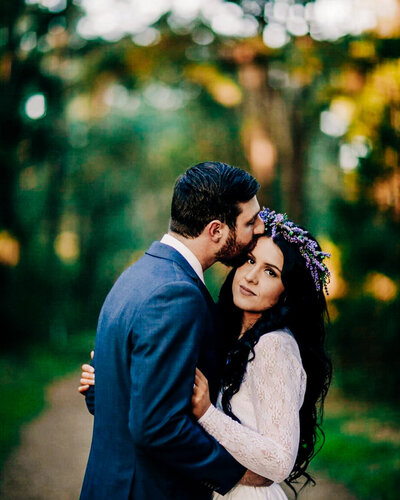 the groom kissing the forehead of the bride as the bride looks in the camera