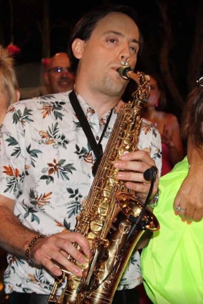 Saxophonist in tropical shirt performing at a vibrant outdoor party with guests dancing nearby.