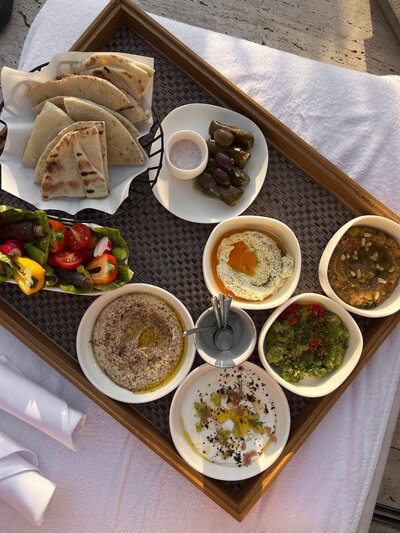 A wooden tray with dishes including hummus, baba ghanoush, tabbouleh, stuffed grape leaves, and pita bread. Accompanied by a salad with cherry tomatoes and olives, and a small bowl of sauce. Sunlight casts soft shadows on the table.