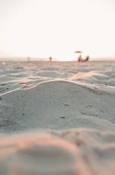 A photograph taken on the beach low down looking along the sand with people and umbrellas in the hazy distance.