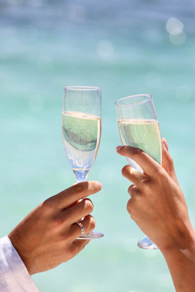 Two hands toasting with champagne glasses against a bright blue ocean background.