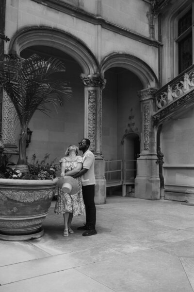 A black and white vintage photograph of a couple sharing a romantic embrace in a garden courtyard. The man has dark skin and wears a white shirt; the woman has light skin and wears a patterned dress. They are standing near a large urn and Mediterranean-style arches.