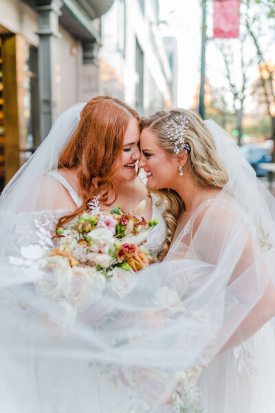 Two brides sharing a joyful moment under a veil, holding romantic pastel bouquets at their Austin wedding, planned by Eventive Designs.