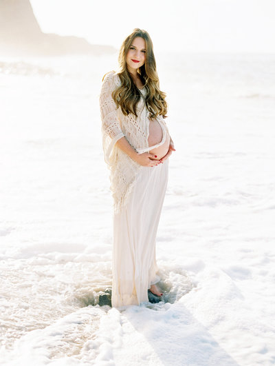Photograph of a maternity session on the beach in Ventura County where a pregnant woman smiles at the camera in the surf