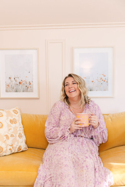 Mollie Mason in a purple dress smiling sitting on a yellow couch holding a coffee mug