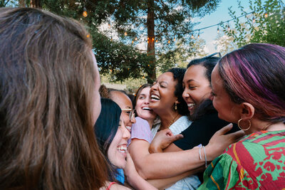 A group of multi-cultural women are gathered in a tight, joyful embrace around a bride to be at her outdoor bridal shower. They are all smiling and laughing, surrounded by a warm atmosphere created by string lights hanging above with trees and bright blue sky in the background.