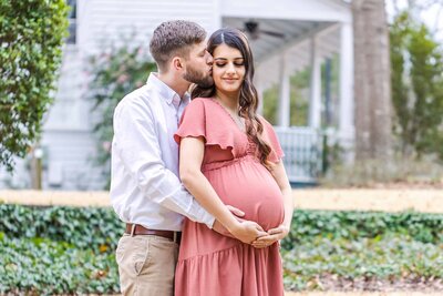 A husband kissing his wife on the cheek while they rest their hands on her baby bump in Lexington SC