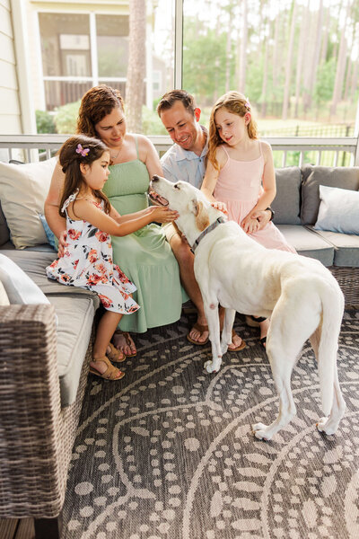 A family of four sits on an outdoor sofa petting a large white dog, captured by a talented family photographer in Fayetteville, NC. The mother in a green dress, father in a light shirt, and two young girls in floral and pink dresses are smiling against the backdrop of trees on the patio.