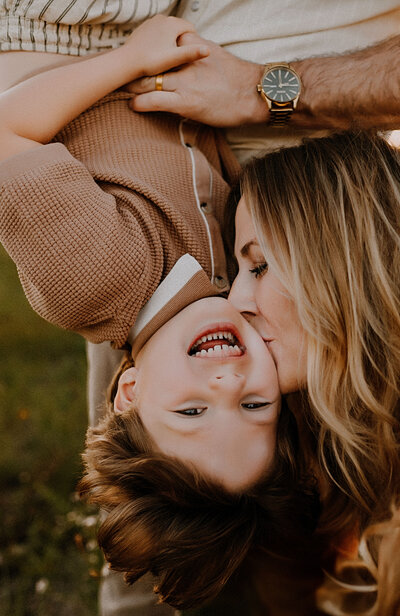 A little boy hangs upside down and laughs as his mom kisses him on the cheek in fort worth, texas