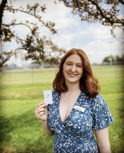 Redheaded woman wearing a blue floral dress standing in a green field wearing a name tag and holding a business card