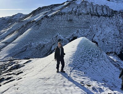 A photographer stands on top of a snowy glacier with a snow topped mountain behind her. She is smiling at the camera while holding a camera in her hand.