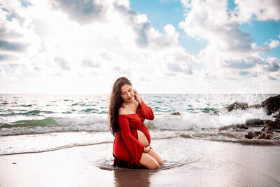 woman wearing a red maternity dress sitting along the edge of the water at the beach
