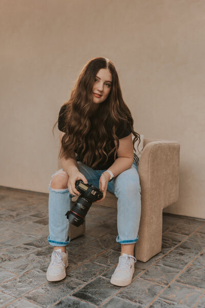 Hannah Burdett from Hannah Grace Photo sitting on a tan chair, holding her camera and softly smiling. Behind her is a tan wall the same color as the chair and weathered stone bricks for the ground.