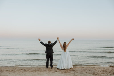 bride and groom holding hands in the air and celebrating on a beach. photo taken by a Wisconsin Elopement Photographer