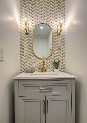 Elegant bathroom upgrade featuring a custom gray vanity with marble countertop, gold fixtures, designer herringbone tile accent wall, and modern lighting—combining functionality with high-end design