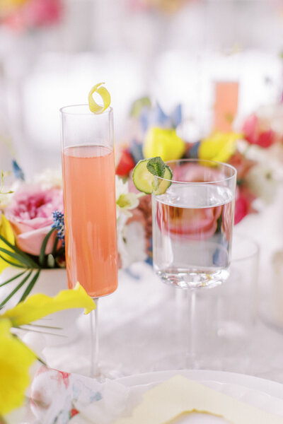 A tall glass of pink drink with a lemon twist and a glass of water with a lime slice sit on a white table adorned with colorful flowers, perfectly capturing the elegant touch of a Canadian wedding planner.