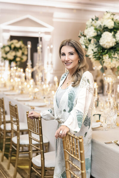 A portrait of Patrice Golesorkhi wearing an elegant floral dress stands smiling beside a beautifully decorated dining table. The table features gold chairs, crystal tableware, and abundant floral arrangements with white roses and greenery. Candles add a warm glow to the setting.