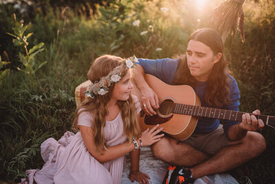 Young girl in a floral crown smiling at her father while he plays the guitar at sunset