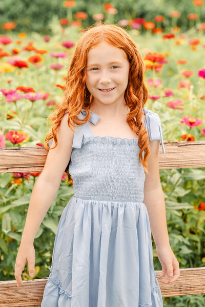 A young red head wearing a blue dress smiles in a wildflower field. Photo taken by Justine Renee Photography in Chesapeake, Virginia.