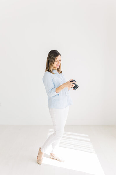 An Austin TX Photographer stands in a white room and looks down at her camera with a smile on her face.