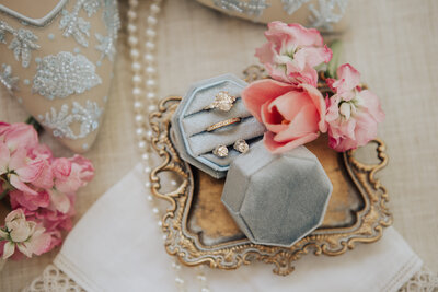 A close up photo of wedding rings in a blue ring box on a gold platter with pink flowers and a string of pearls.
