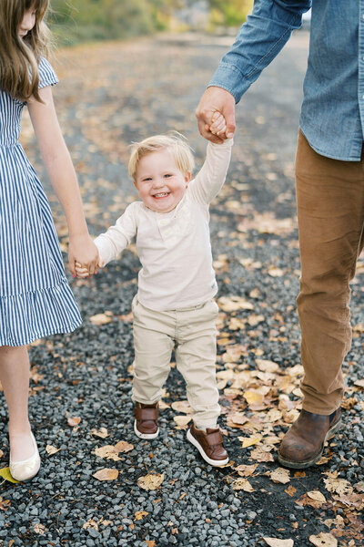 toddler holds hands with family, surrounded by leaves on the ground in fall family photography
