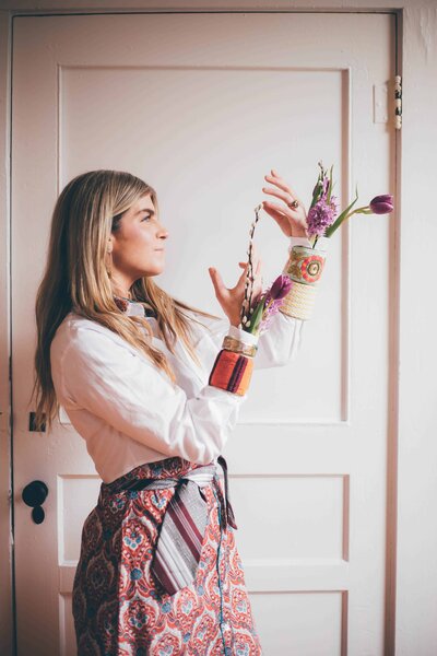 A woman stands against a white wall looking at colorful bracelets on her arms captured by personal branding photographer, Britt Elizabeth Photography.