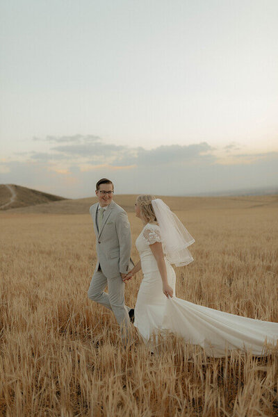 A groom holds the hand of his bride as he looks back at her. They're running through a wheat field together.