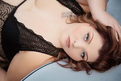 A woman with wavy auburn hair lies on a soft surface, wearing a black lace top. She gazes softly into the camera, conveying serenity and confidence.
