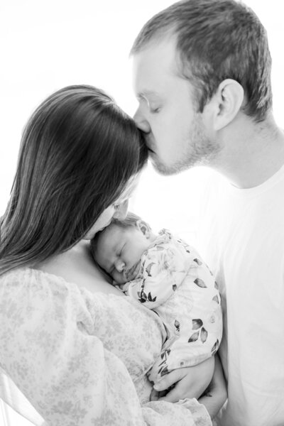 Father kissing wife's forehead while she holds newborn | Newborn Session Photographer