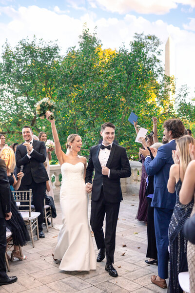 A bride and groom walk down an outdoor aisle, hand in hand. The bride raises her bouquet triumphantly, while guests on either side applaud and smile. A tall monument is visible in the background, surrounded by trees and a partly cloudy sky.