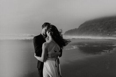 Bride and groom holding each other tight on on the Oregon Coast as the wind whips her long hair 