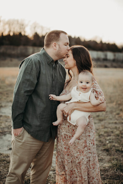 Family photo with dad kissing mom’s forehead while baby smiles brightly.