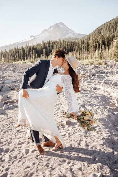 The bride is standing at the base of mountains as she kicks up her leg. She is wearing combat boots and a wedding dress. She has a hiking backpack on with flowers in it. She is smiling towards the ground with one arm lifted in the air. The grass is brown and hte mountains are blue.