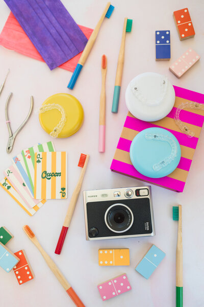 Flatlay of vintage film camera, colorful wooden toothbrushes, striped playing cards, colored dice, and assorted orthodontic tools