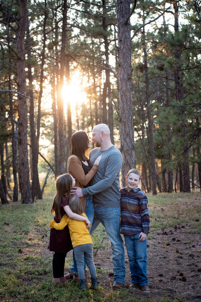 Mom and dad kiss while their three toddlers play around them looking on as posed by a Denver family photographer