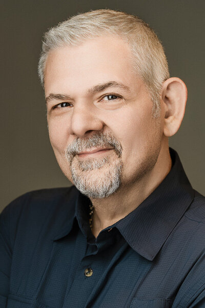 Professional headshot of wedding photographer Alex Kaplan, smiling warmly with gray hair and beard, wearing a navy shirt against a soft background.