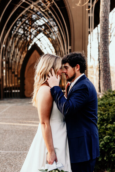 The groom caresses his brides face in front of the glass chapel leaning in for a kiss