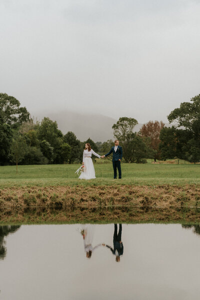 An artistic wedding photo of a Coffs Harbour couple standing against a misty mountain backdrop. They are mirrored in the reflection of the lake before them.