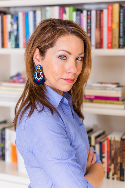 woman writer standing in blue shirt crossing harms in front of her library full of books