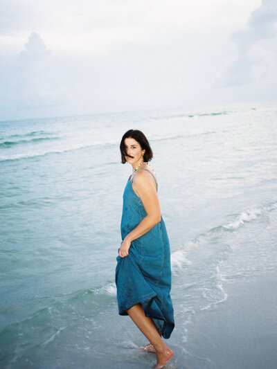 Courtney Leigh in a blue dress walking in in the water along the beach