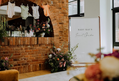 Floral baby shower setup with a sign reading ‘Baby Kai’s Secret Garden,’ colorful blooms, and baby clothes hanging on display, styled by Yours Truly, a wedding planner and designer in New Jersey.