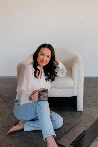 A successful brand and Showit design partner smiles confidently while sitting on the floor with her coffee mug and laptop. Wearing a white blouse and light blue jeans, she represents one of the established designers who transformed her business by incorporating white label design services after completing The White Label Way course, creating additional revenue streams while maintaining her creative freedom.