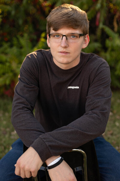 A high school senior boy with glasses sits outdoors, wearing a dark Patagonia sweater. The soft, golden sunlight highlights his facial features and creates a warm, natural look, making this portrait perfect for showcasing his personality and confidence.