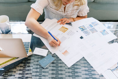 Blonde woman sits on geometric blue and white rug on phone wearing a blue silk blouse, white jeans, and brown sandals