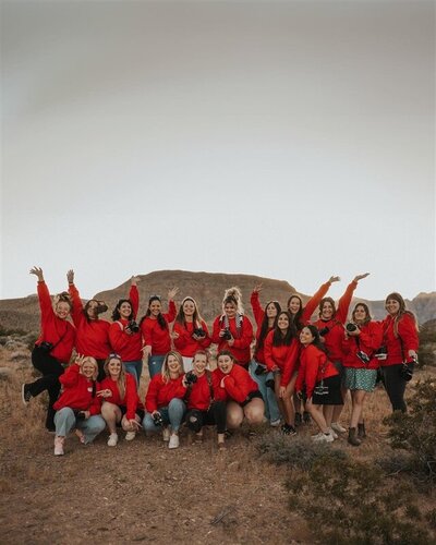 A group of photographers at Aglow, all wearing red crewneck sweatshirts, pose together in a desert landscape, their arms raised in excitement.